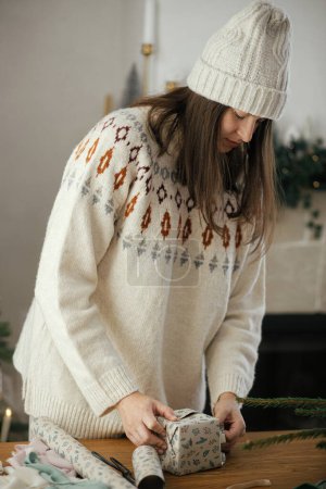 Foto de Mujer en suéter acogedor y sombrero envolviendo regalo de Navidad en papel sobre mesa de madera con decoraciones festivas en habitación escandinava decorada. ¡Feliz Navidad! Manos regalo de embalaje - Imagen libre de derechos