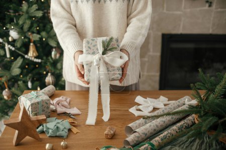 Téléchargez les photos : Femme tenant cadeau de Noël décoré élégant près sur le fond de la table avec des décorations festives dans la chambre scandinave moderne. Joyeux Noël et joyeuses fêtes! - en image libre de droit