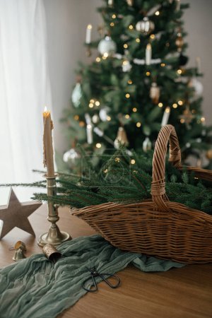 Foto de Elegante cesta rústica con ramas de abeto, vela vintage y estrella de madera en la mesa contra el árbol de Navidad decorado festivo en la habitación escandinava. Feliz Navidad y Felices Fiestas! - Imagen libre de derechos