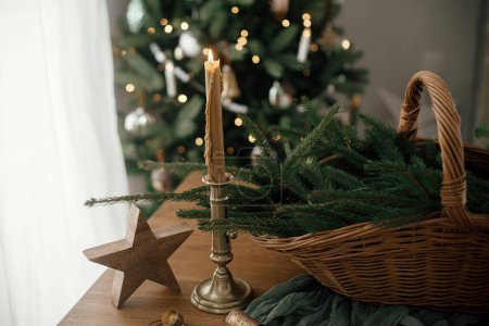 Téléchargez les photos : Joyeux Noël et Joyeuses fêtes ! Panier rustique élégant avec branches de sapin, bougie vintage et étoile en bois sur la table contre l'arbre de Noël décoré de fête dans la chambre scandinave. - en image libre de droit