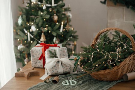 Photo for Stylish wrapped christmas gifts, rustic basket with fir branches and modern decorations against festive decorated tree in scandinavian room. Merry Christmas and Happy holidays! - Royalty Free Image