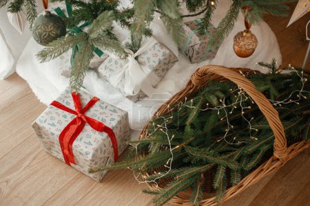 Photo for Stylish wrapped christmas gifts and rustic basket with fir branches under festive decorated christmas tree in scandinavian room. Merry Christmas and Happy holidays! - Royalty Free Image