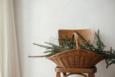 Foto de Elegante cesta rústica con ramas de abeto y luces en silla de madera contra la pared rural en habitación escandinava. Feliz Navidad y Felices Fiestas! Navidad naturaleza muerta rústica - Imagen libre de derechos