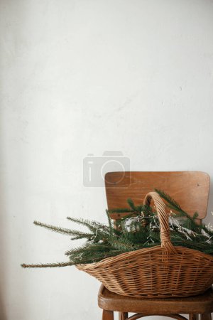 Foto de Elegante cesta rústica con ramas de abeto y luces en silla de madera contra la pared rural en habitación escandinava. Feliz Navidad y Felices Fiestas! Navidad naturaleza muerta rústica - Imagen libre de derechos