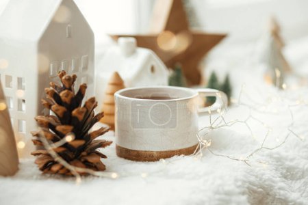 Foto de Acogedor invierno naturaleza muerta. Elegante taza de té con decoración moderna de Navidad, cono de pino, estrella de madera y árbol, luces doradas sobre una suave manta caliente en el alféizar de la ventana. Feliz Navidad y Felices Fiestas - Imagen libre de derechos