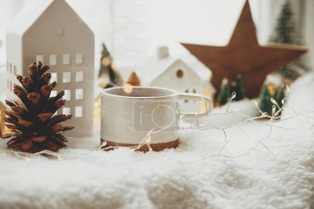 Photo for Winter hygge. Stylish cup of tea with modern cute christmas houses, pine cone, wooden star and tree, golden lights on soft warm blanket on windowsill. Christmas cozy still life. Merry Christmas! - Royalty Free Image