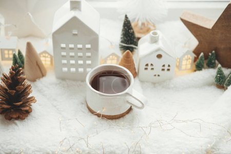Photo for Stylish cup of tea with modern christmas houses, pine cone, wooden star and tree decor, golden lights on warm blanket on windowsill. Cozy Christmas, scandinavian style. Christmas background - Royalty Free Image