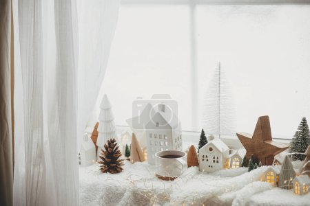 Téléchargez les photos : Élégante tasse de thé avec des maisons de Noël modernes, cône de pin, étoile en bois et décor d'arbre, lumières dorées sur couverture chaude sur le rebord de la fenêtre. Noël douillet, style scandinave. Fond de Noël - en image libre de droit