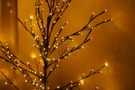 Foto de Elegante árbol minimalista brillante en la habitación acogedora noche. Decoración de Navidad escandinava, mínimo moderno árbol negro ramas luces doradas bokeh, espacio de copia. ¡Felices fiestas! Iluminación festiva - Imagen libre de derechos