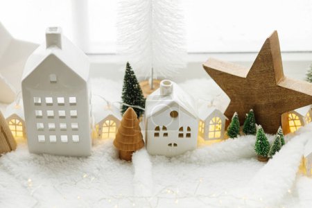 Téléchargez les photos : Village d'hiver miniature atmosphérique. Élégantes petites maisons en céramique et des arbres de Noël en bois sur une couverture de neige molle avec des lumières incandescentes. Noël fond blanc moderne. Joyeuses fêtes! - en image libre de droit