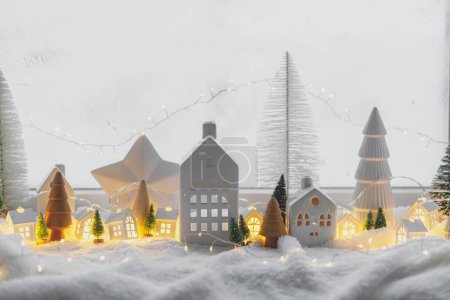 Photo for Atmospheric miniature winter village. Stylish little ceramic houses and christmas wooden trees on snow blanket with glowing lights in evening. Christmas modern background. Happy Holidays! - Royalty Free Image