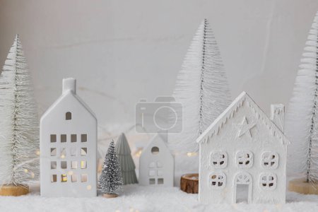Photo for Cozy christmas miniature village. Stylish little ceramic houses and trees on snow blanket with golden lights on white background. Atmospheric winter village  modern still life. Merry Christmas! - Royalty Free Image