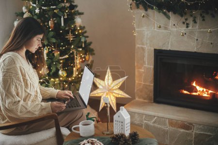 Photo for Woman in cozy sweater holding credit card and typing on laptop with blank screen in festive decorated christmas room with lights. Christmas shopping online and black friday sales - Royalty Free Image