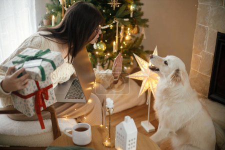 Photo for Happy woman holding stylish christmas gifts with credit card and playing with cute white dog in festive decorated christmas room with lights. Christmas shopping online and black friday sales. - Royalty Free Image