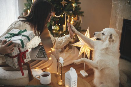 Téléchargez les photos : Femme heureuse tenant des cadeaux de Noël élégants avec carte de crédit et jouant avec chien blanc mignon dans la salle de Noël décorée festive avec des lumières. Achats de Noël en ligne et ventes du vendredi noir. - en image libre de droit