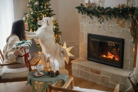 Foto de Mujer feliz sosteniendo elegantes regalos de Navidad con tarjeta de crédito y jugando con lindo perro blanco en la sala de Navidad decorada festiva con luces. Compras de Navidad en línea y negro viernes ventas. - Imagen libre de derechos
