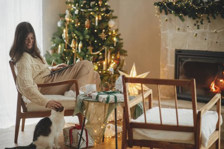 Foto de Mujer feliz trabajando en el ordenador portátil y acariciando lindo gato en la sala de Navidad decorada festiva con luces de árbol. Vacaciones de Navidad y mascotas. Trabajo remoto en vacaciones de invierno - Imagen libre de derechos