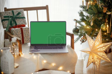 Photo for Laptop with empty screen and stylish christmas gifts on modern chair on background of festive decorated tree, golden lights and star in room. Christmas shopping online and sales. Laptop mock up - Royalty Free Image