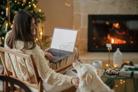 Téléchargez les photos : Femme heureuse travaillant sur ordinateur portable et caressant chien blanc mignon dans la salle de Noël décorée festive avec des lumières. Vacances de Noël et animaux domestiques. Travail à distance pendant les vacances d'hiver - en image libre de droit