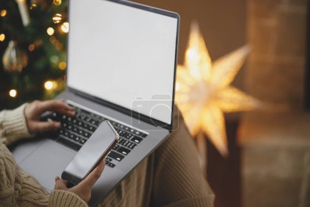 Photo for Woman in cozy sweater working on laptop with empty screen and holding phone on background of christmas tree lights in stylish festive decorated room. Christmas shopping online and remote work - Royalty Free Image