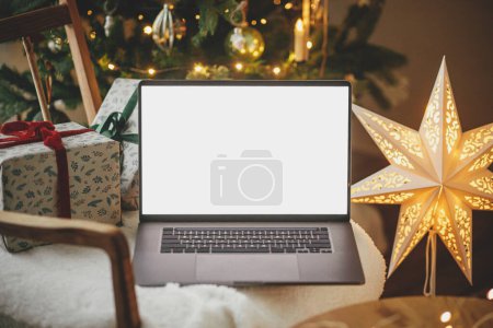 Téléchargez les photos : Ordinateur portable avec écran vide et cadeaux de Noël élégants sur chaise moderne sur fond d'arbre décoré de fête, lumières dorées et étoile dans la chambre. Achats de Noël en ligne et les ventes. Ordinateur portable mock up - en image libre de droit