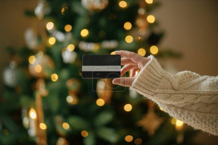 Téléchargez les photos : Achats de Noël en ligne et ventes du vendredi noir. Main dans un pull confortable tenant la carte de crédit près sur le fond des lumières de l'arbre de Noël dans la chambre décorée festive - en image libre de droit