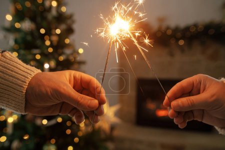 Téléchargez les photos : Brûlant scintillements dans les mains sur fond de cheminée de campagne moderne et arbre de Noël avec des lumières dorées. Bonne année ! Feux d'artifice dans les mains, couple célébrant dans la salle décorée festive - en image libre de droit