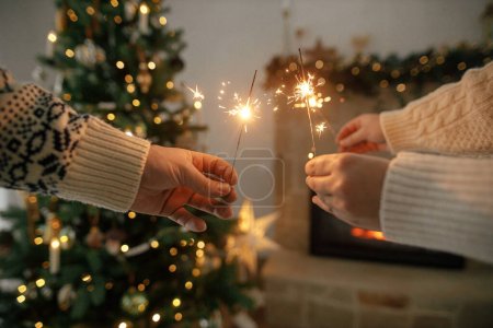 Foto de ¡Feliz Año Nuevo! Manos que sostienen fuegos artificiales ardientes contra la chimenea moderna y el árbol de Navidad con luces doradas. Amigos y familiares celebrando con bengalas ardientes en las manos, víspera atmosférica - Imagen libre de derechos