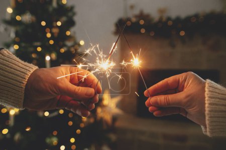 Photo for Hands holding burning fireworks against modern fireplace and christmas tree with golden lights. Happy New Year! Friends and family celebrating with burning sparklers in hands, atmospheric eve - Royalty Free Image
