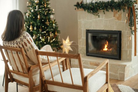 Foto de Mujer en suéter acogedor relajarse en la silla moderna y mirando a la chimenea con manto festivo en el fondo de elegante árbol de Navidad decorado con luces. Vacaciones de invierno - Imagen libre de derechos
