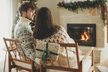 Photo for Happy couple in cozy sweaters exchanging stylish wrapped christmas gifts on background of fireplace with festive mantle and modern decorated christmas tree with lights. Merry Christmas! - Royalty Free Image