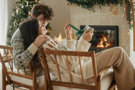 Photo for Happy young family in cozy sweaters opening stylish christmas gifts on background of fireplace with modern festive mantle and christmas tree with lights. Happy Holidays! - Royalty Free Image