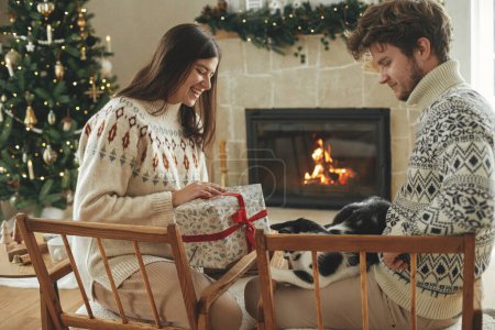 Photo for Happy couple in cozy sweaters playing with cat and exchanging stylish christmas gifts on background of fireplace with festive mantle and modern christmas tree with lights. Merry Christmas! - Royalty Free Image