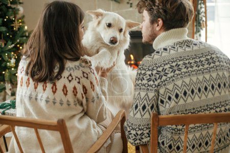 Téléchargez les photos : Joyeux jeune famille dans des pulls confortables étreignant avec chien mignon et relaxant sur fond de cheminée et arbre de Noël moderne. Joyeux Noël ! Animaux domestiques et vacances d'hiver - en image libre de droit