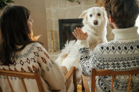 Photo for Happy young family in cozy sweaters playing with cute dog and relaxing on background of fireplace and modern christmas tree. Merry Christmas! Pets and winter holidays - Royalty Free Image