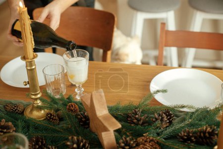 Foto de Cena festiva de Navidad. Manos vertiendo champán en copas sobre mesa de madera con ramas de abeto y decoración de conos de pino, vasos y cubiertos. ¡Feliz año nuevo! Acuerdo de vacaciones - Imagen libre de derechos