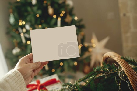 Foto de Mano sosteniendo la tarjeta de felicitación vacía en el fondo de elegantes regalos envueltos en Navidad, ramas de abeto y árbol festivo con luces doradas. La tarjeta de Navidad se burla. Espacio para texto - Imagen libre de derechos