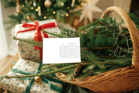 Photo for Christmas card mock up. Empty greeting card on background of stylish christmas wrapped gifts, fir branches in rustic basket and festive tree with golden lights. Space for text. - Royalty Free Image