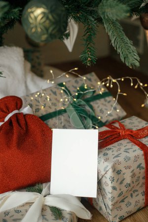 Téléchargez les photos : Carte de Noël maquillée. Carte de vœux vide sur fond de cadeaux de Noël enveloppés élégants sous l'arbre de Noël festif avec des lumières dorées. Espace pour le texte. Modèle de salutations de saison - en image libre de droit