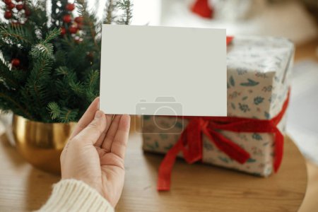 Téléchargez les photos : Carte de Noël maquillée. Main tenant carte de vœux vide sur élégant cadeau de Noël enveloppé et branches de sapin avec des lumières de Noël festives sur table en bois. Espace pour le texte. Carte postale de voeux de saison - en image libre de droit