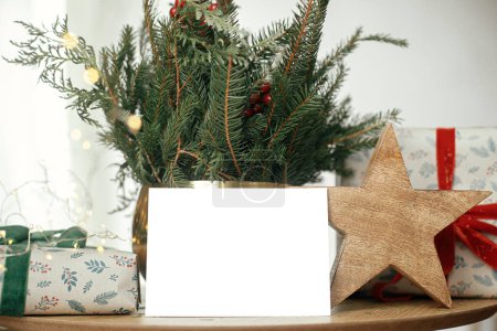 Téléchargez les photos : Carte de vœux vide, élégant cadeau de Noël enveloppé, branches rustiques en étoile et sapin avec des lumières sur une table en bois. Carte de Noël maquillée. Espace pour le texte. Modèle de carte postale de voeux de saison - en image libre de droit