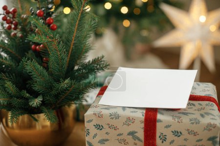 Téléchargez les photos : Carte de Noël maquillée. Carte de voeux vide cadeau de Noël enveloppé élégant contre l'arbre de Noël festif avec des lumières dorées. Espace pour le texte. Modèle de carte postale de voeux de saison - en image libre de droit