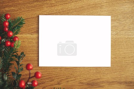 Téléchargez les photos : Carte de Noël maquette à plat. Carte de vœux vide et branches de sapin avec des baies rouges sur table en bois. Espace pour le texte. Modèle de carte postale de voeux de saison - en image libre de droit