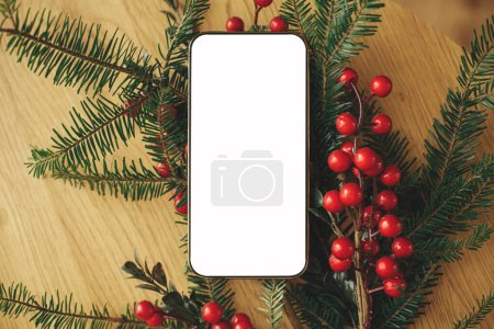 Téléchargez les photos : Téléphone de Noël se maquiller à plat. Téléphone écran vide et branches de sapin avec des baies rouges sur table en bois rustique. Espace pour le texte. Publicité de Noël, modèle d'application - en image libre de droit