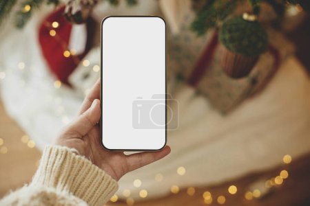 Foto de Smartphone de mano con pantalla vacía contra elegantes regalos navideños y luces doradas. El teléfono de Navidad se burla. Espacio para el texto. Publicidad de Navidad, plantilla de aplicación smartphone - Imagen libre de derechos