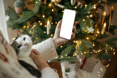 Foto de Mano sosteniendo smartphone con pantalla vacía y abrazando lindo gato contra elegante árbol de Navidad festivo con luces doradas. El teléfono de Navidad se burla. Espacio para el texto. Plantilla de aplicación de publicidad Navidad - Imagen libre de derechos