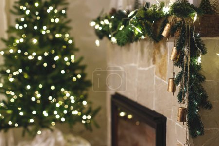Photo for Atmospheric christmas eve. Stylish decorated christmas tree with lights  and modern decor on fireplace mantel with bells. Modern christmas scandinavian living room. - Royalty Free Image