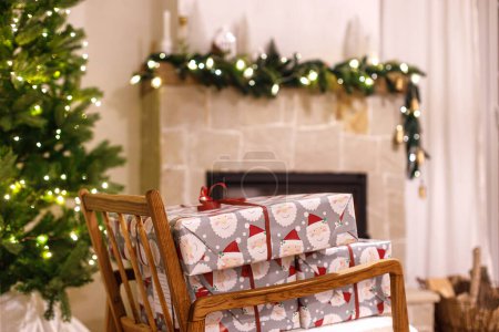 Téléchargez les photos : Joyeux Noël ! Cadeaux de Noël enveloppés élégants sur fauteuil contre l'arbre de Noël avec des lumières festives et cheminée à combustion confortable. Veille de Noël atmosphérique, temps des fêtes. - en image libre de droit