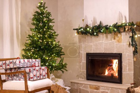 Foto de Elegantes regalos de Navidad envueltos en sillón contra el árbol de Navidad con luces festivas y acogedora chimenea. Nochebuena, tiempo de vacaciones. Sala de estar atmosférica con regalos de Navidad - Imagen libre de derechos