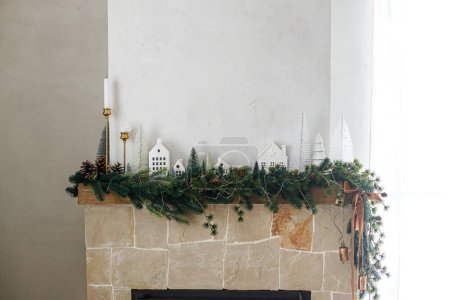 Téléchargez les photos : Élégante cheminée décorée de Noël. Arbres de Noël modernes, maisons, cônes de pin et branches d'épinette sur cheminée manteau avec cloches en laiton et ruban. Décor scandinave de salon de Noël - en image libre de droit